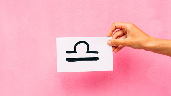 A hand hold a paper with a Libra sign against a pink background. 