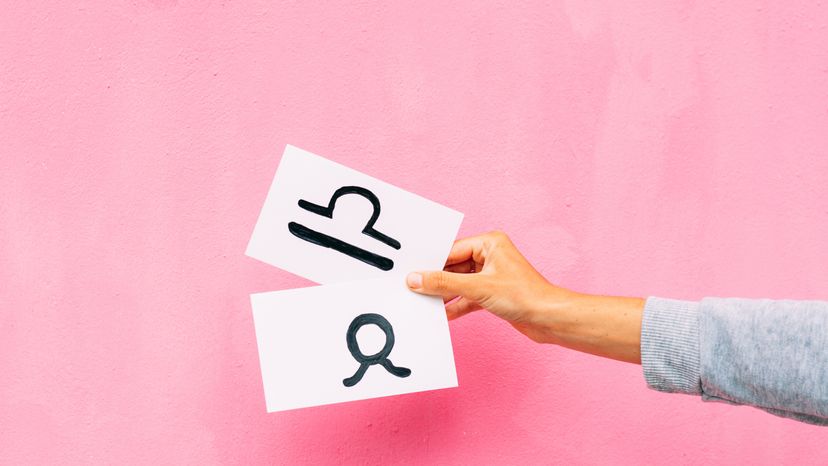 Hand holding a white paper with Libra sign drawn on it, against a pink background. 