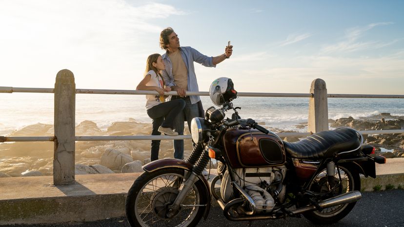 A father taking a picture with his daughter, with a classic motorcycle parked behind them. 