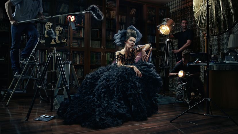 A woman wearing an old dress, sitting on a chair in a movie set.