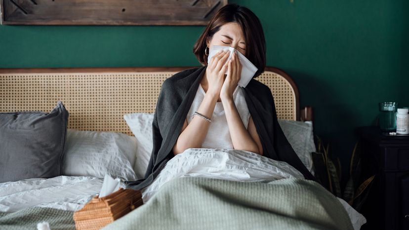 A young Asian woman in bed, blowing her nose with tissue. 