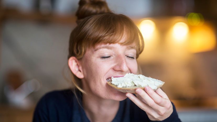 A woman biting into her toast, topped with cream cheese. 