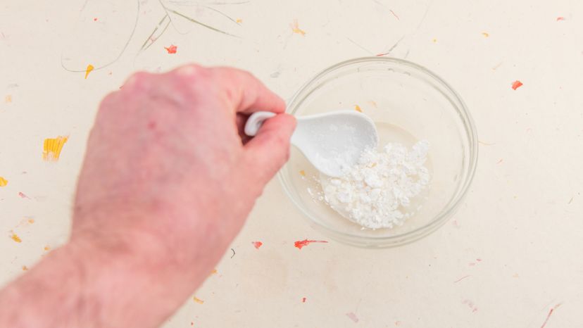 A human hand using a white spoon to stir cornstarch and water.