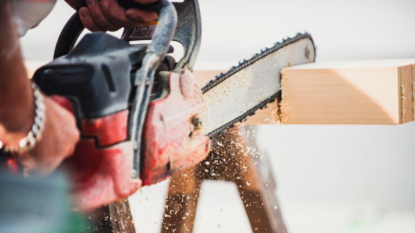 A close up image of a man sawing a wooden beam with a chain saw. 