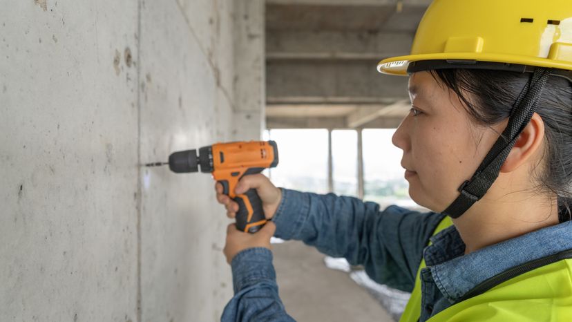 一个亚洲女性,建筑工人用电钻在墙上钻洞。”width=