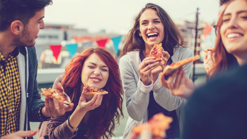 A group of friends eating pizza outdoors and laughing. 