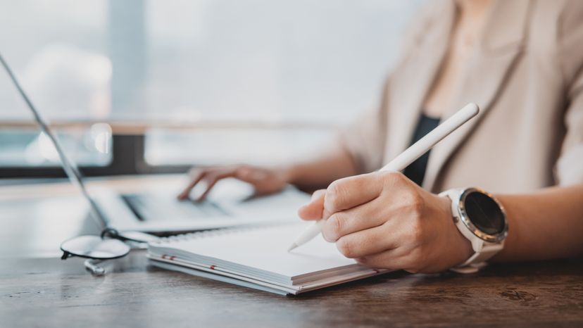 A woman writing on a notepad while using a laptop.