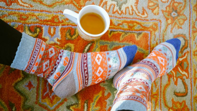 A close up of a woman wearing knitted socks on a rug with a cup of tea nearby. 