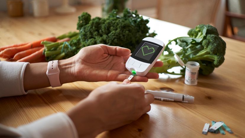 A young woman sitting at a table measuring her blood sugar level with a glucometer, with vegetable scattered on the table. 