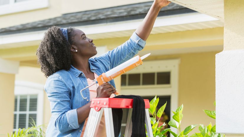 A young woman standing on a ladder outside her house while using a caulking gun to repair a leak.