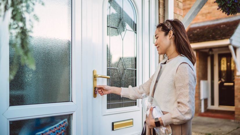 A young woman holding a bottle of water unlocks the front door of her house.