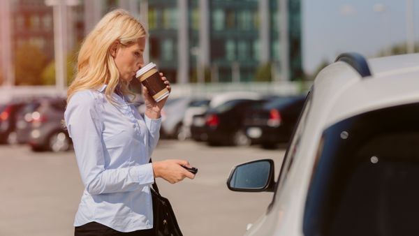 Businesswoman talking on the phone outdoors and standing in front of the car, drinking coffee, and unlocking the vehicle.
