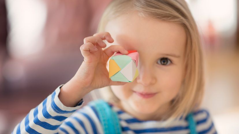 A little girl holding up a cube made out of paper. 