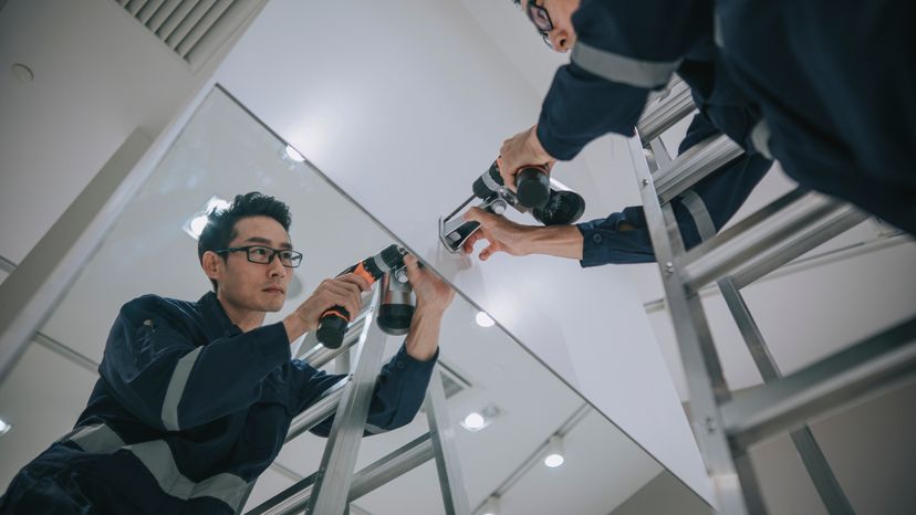 An Asian man on a ladder, installing a CCTV camera at a store. 