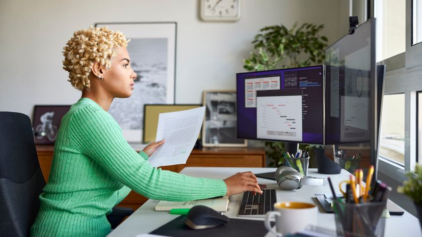 A side view of a businesswoman sitting at a desk, holding a document, while using  a computer. 