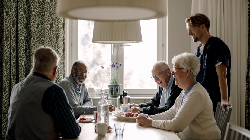 A group of four elderly people seated at a table while their nurse laughs with them. 