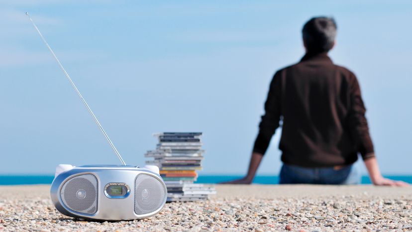 A man sitting at the beach with a stack of CDs and a CD player behind him. 
