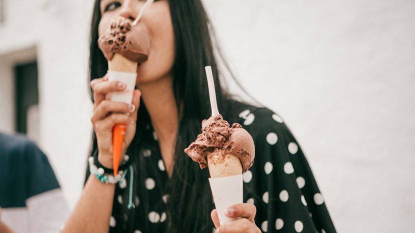 A young lady licking a chocolate ice cream while holding another. 