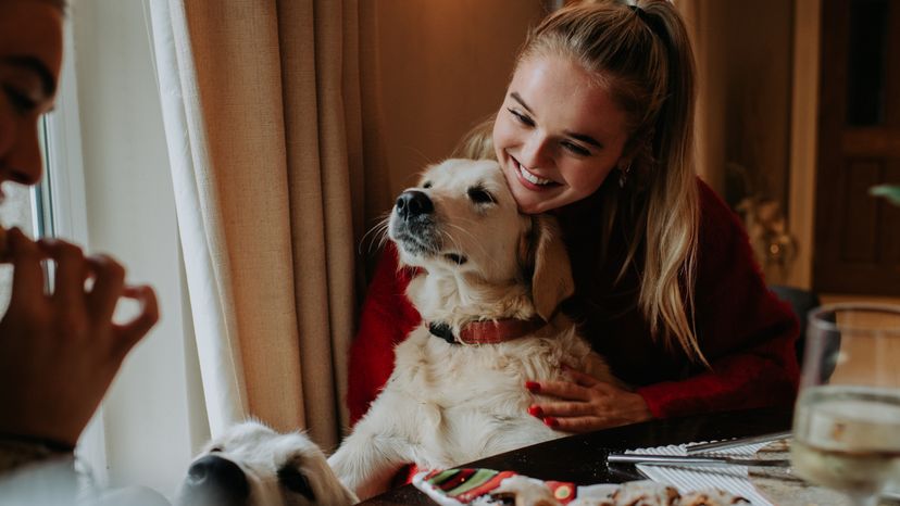 Two young women sitting at a table with a golden retriever. 