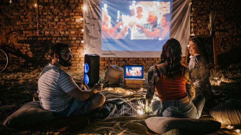 A man and two women sitting outdoors while watching a movie on a projected screen. 