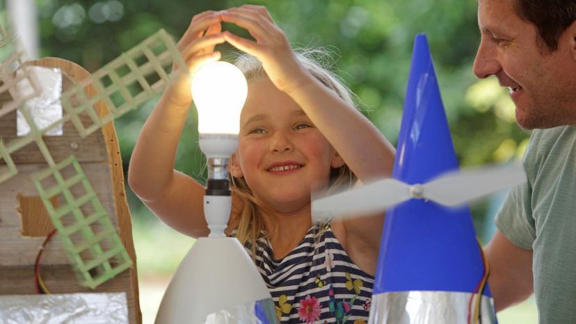 A little girl holding her hand over a lit light bulb, while laughing with her dad. 