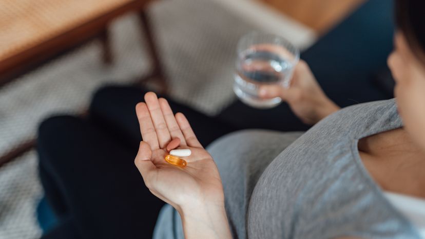 A close up of a pregnant woman holding her prenatal vitamins in her left hand while holding a cup of water in her right hand. 