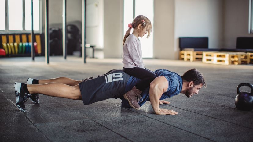 A man in the gym doing push ups with a little girl sitting on his back. 