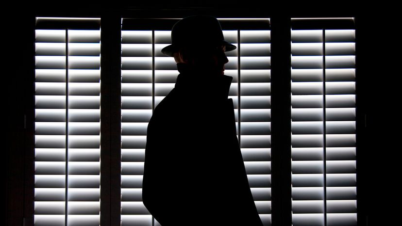 A man in a coat and hat, standing beside a window.