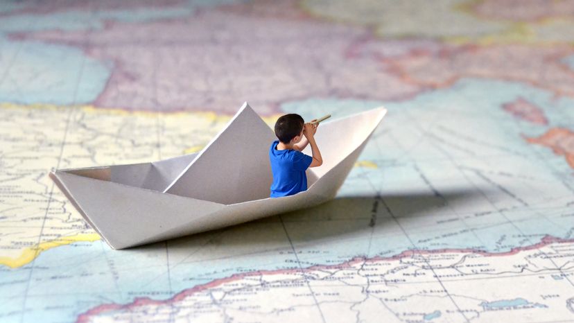 A cartoon picture of a boy sailing in a paper boat across a paper map. 