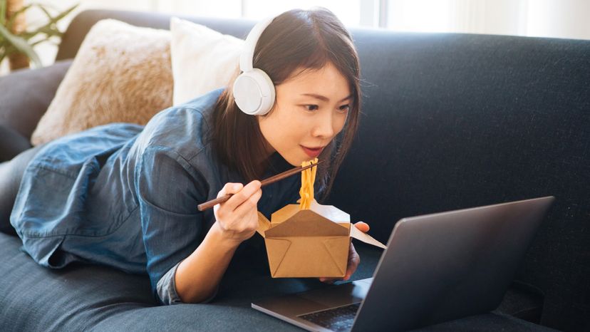 A young woman eating noodles while watching a movie on a laptop. 