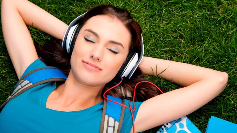 A female college student laying on grass while listening to music on her headphone. 