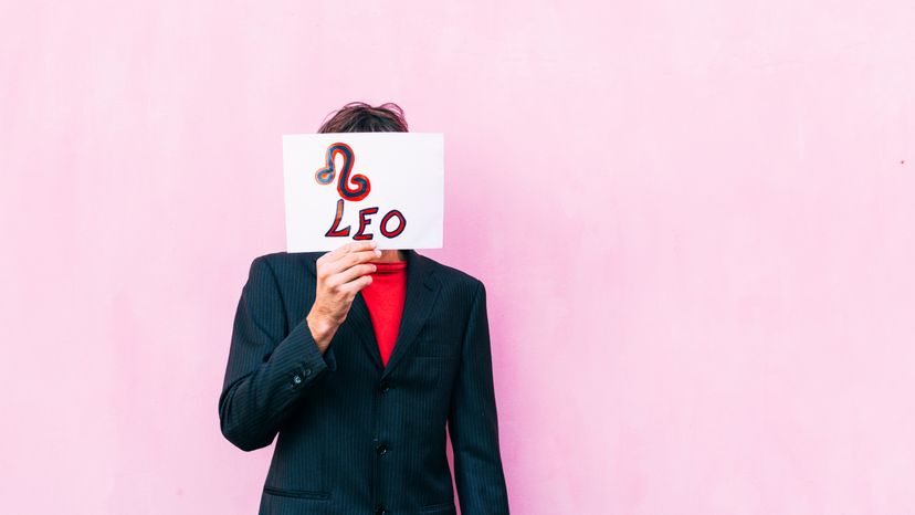 A man holding a white paper with a zodiac sign over his face on a pink background. 