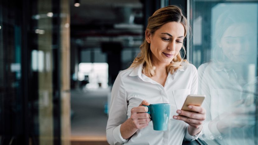 A business woman texting on her cell phone while holding a mug.