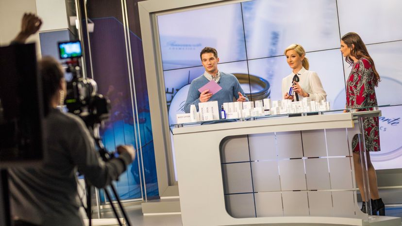 A man and two women standing near a counter full of cosmetic products while being filmed. 