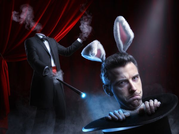 A magician's head pops out of a top hat, wearing rabbit ears; his headless body is in the background holding a wand. Something has gone wrong.