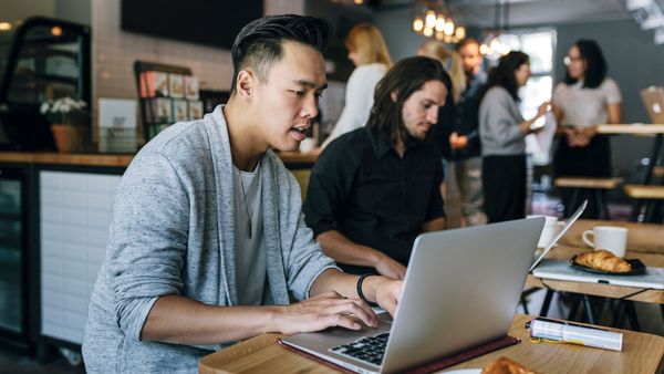 Two young colleagues sitting in a busy business cafe and working on their laptops as they eat.