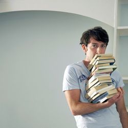 A young man stands holding a large stack of books.