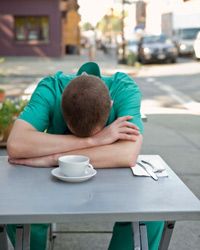 A man in scrubs with his head down on an outdoor café table.