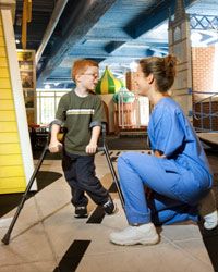 A woman in scrubs kneels to talk with a boy using crutches in a kids' physical therapy center.