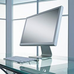 A silver computer monitor sitting on a glass desk.&nbsp;