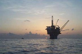 A spar production platform floats at sunset in the Gulf of Mexico. The structure's massive cylindrical hull extends down into the depths for hundreds of feet.