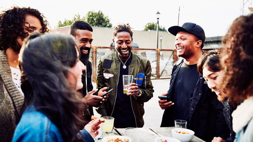 A group of friends stand around a table, laughing, socially drinking alcohol.