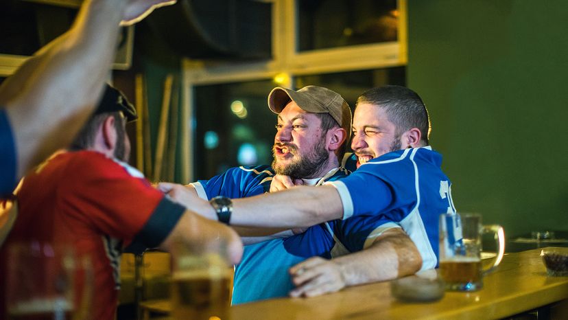 A fight breaks out between two males wearing sports jerseys, while another man tries to break it up.