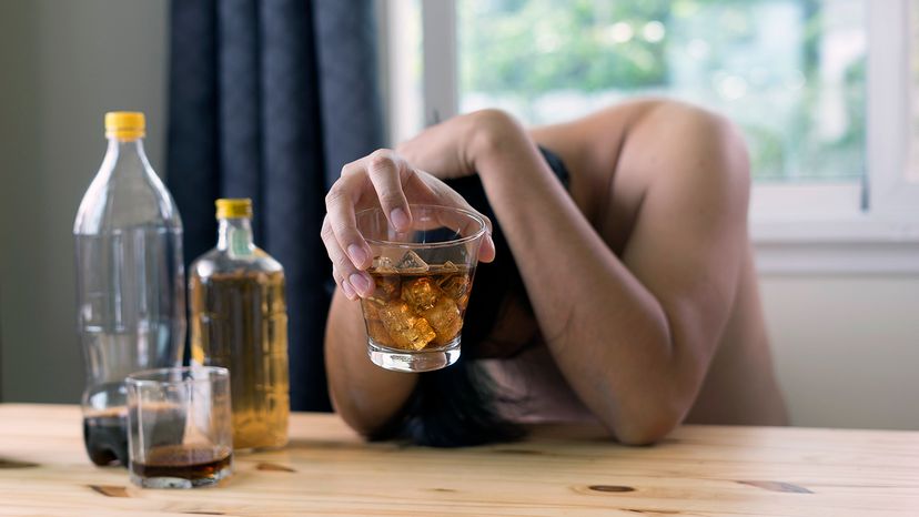 A woman holding a mixed drink is holding her head while sitting at a table.