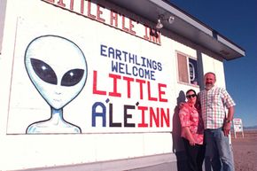 A man and woman stand in front of a mural that says "Earthlings Welcome" and "Little&nbsp;A'Le'Inn".