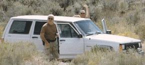 Two men wearing brown shirts and camo pants stand outside of a white SUV.