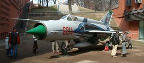 The white, green and red MiG-21 plane, with groups of people standing around, looking at it.