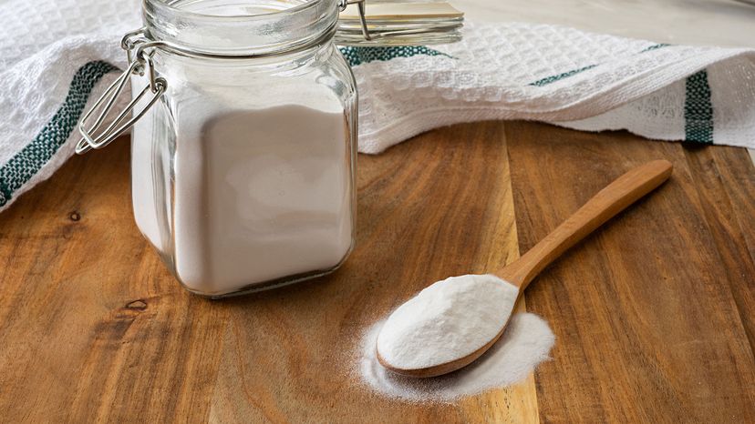 Open jar of baking soda with a wooden spoon of baking soda on a wooden table.