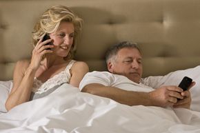 A couple both relax in bed, looking at electronic devices.