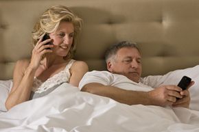 A couple both relax in bed, looking at electronic devices.